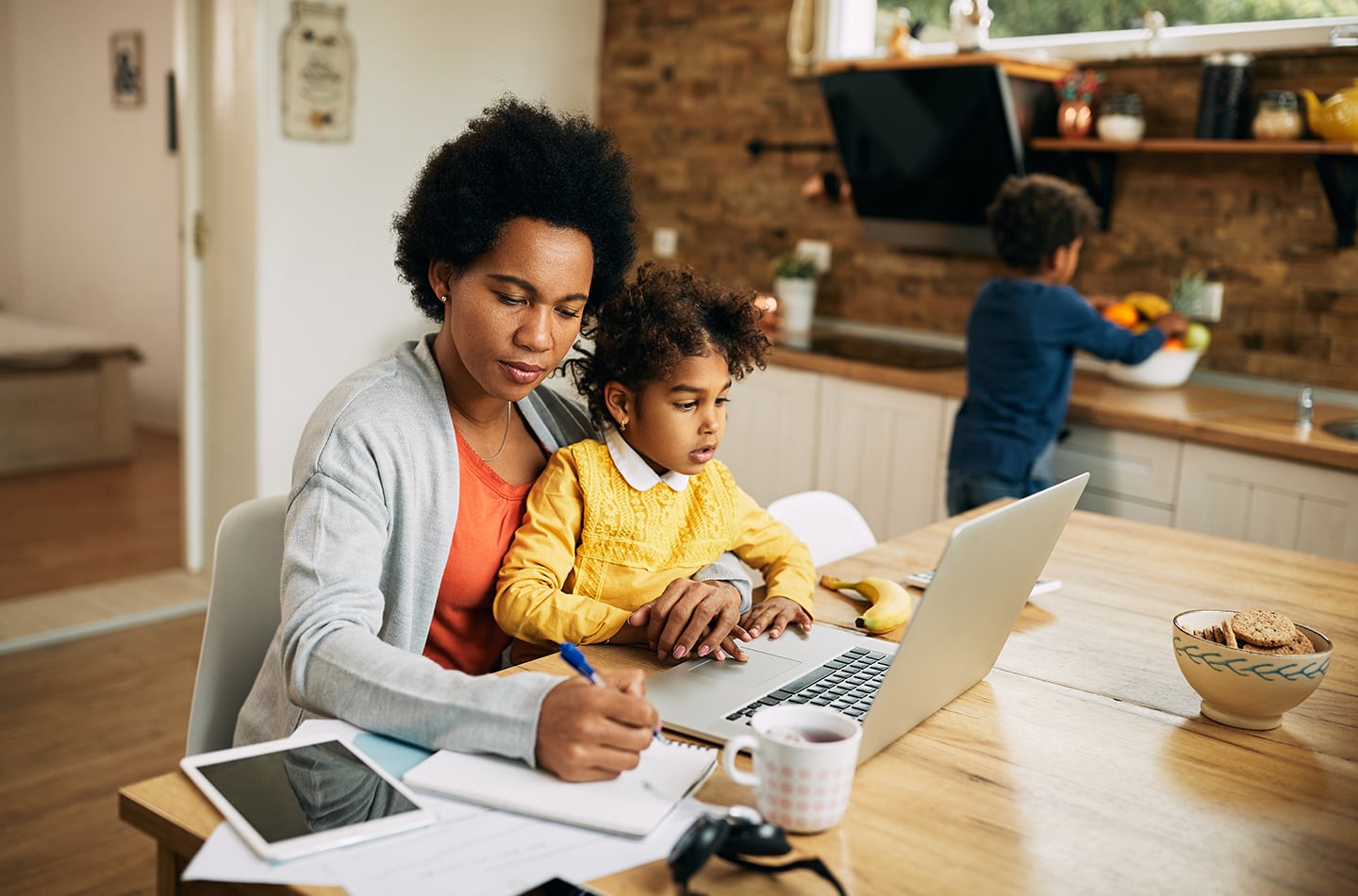 A mother studying with a notebook and laptop at the dinner table with her young child sitting in her lap while an older child cleans fruit behind her