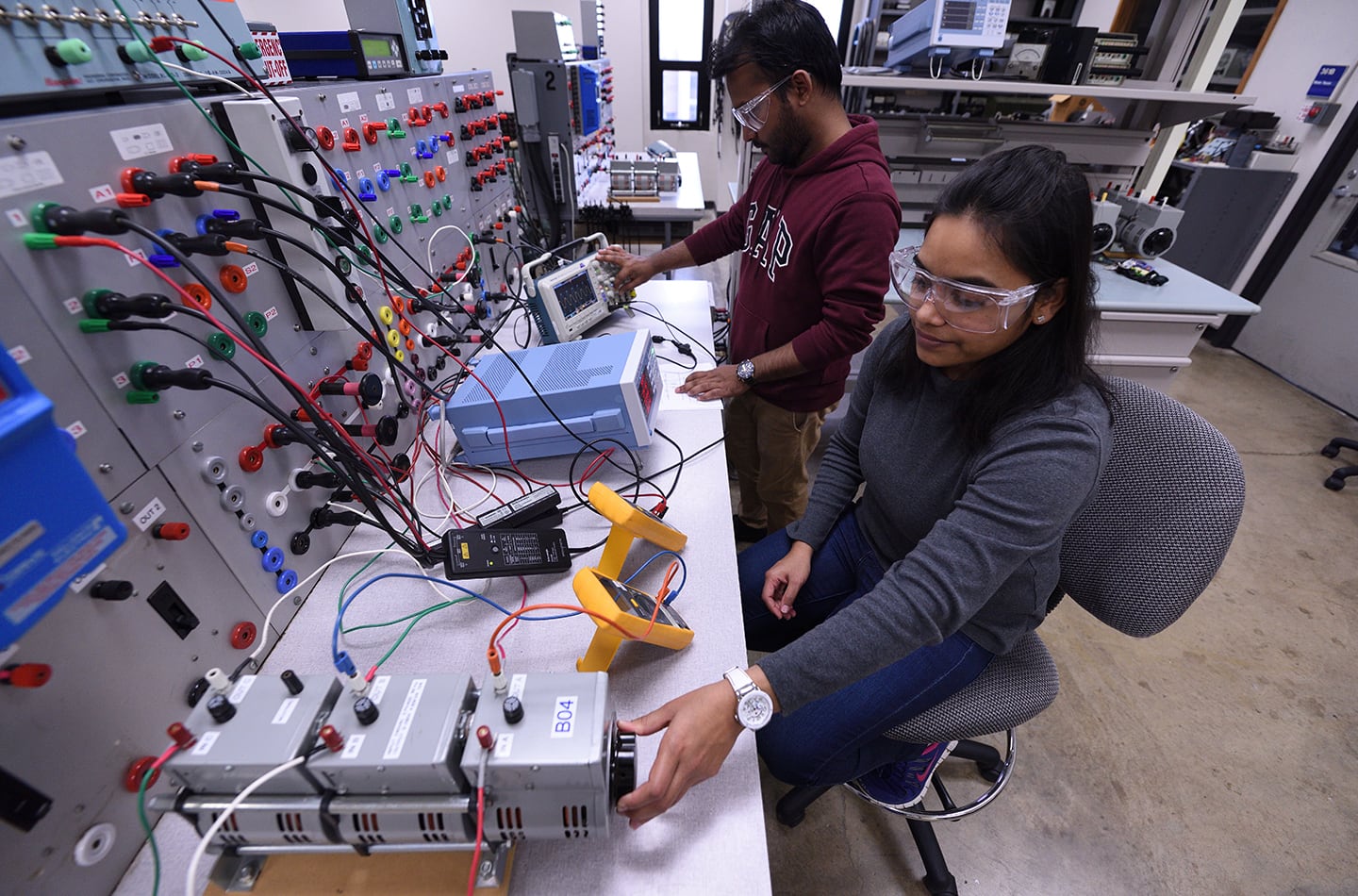 Two engineering students wearing safety glasses work in an electrical lab. One student sits and adjusts a piece of machinery while the other stands nearby, using an oscilloscope and monitoring a complex circuit board with various colorful wires and connectors.