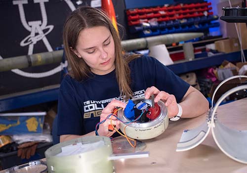 A young woman works on assembling electronic components inside a round metal device at a workshop table, surrounded by tools and equipment.