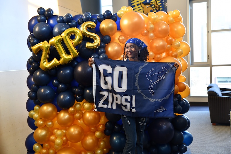 A person stands in front of a balloon display holding a blue flag that reads GO ZIPS! with a kangaroo logo and The University of Akron. The balloons are blue, gold, and orange, with gold letter balloons spelling ZIPS.