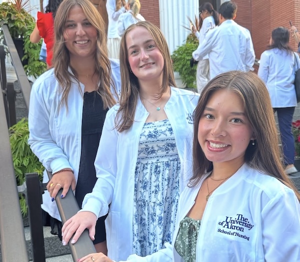 Three smiling women wearing white coats, one with The University of Akron School of Nursing on it, stand on outdoor steps with other people in similar attire in the background.