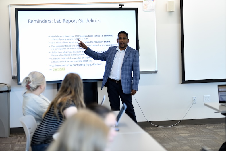 A professor in a plaid blazer gestures toward a projected slide titled 'Reminders: Lab Report Guidelines' while teaching a college classroom of seated students.