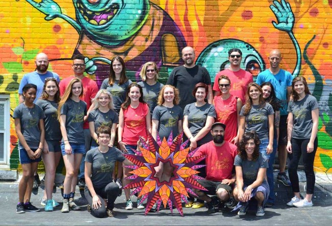 A group of approximately 20 Arts LIFT participants, including university students, professional artists and community partners, pose in front of a brightly colored mural featuring whimsical blue characters. In the foreground, the group displays a vibrant, star-shaped sculptural artwork created as part of the community arts program. The mural and sculpture highlight the collaborative, public art focus of the Arts LIFT apprenticeship.