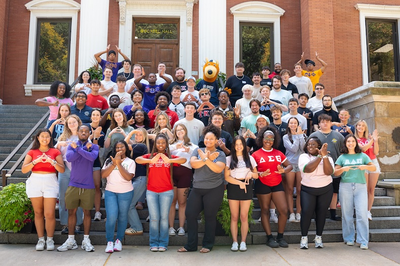 A large group of college students pose on outdoor steps, many wearing Greek letter shirts and making hand symbols, in front of a brick building with columns and a wooden door.