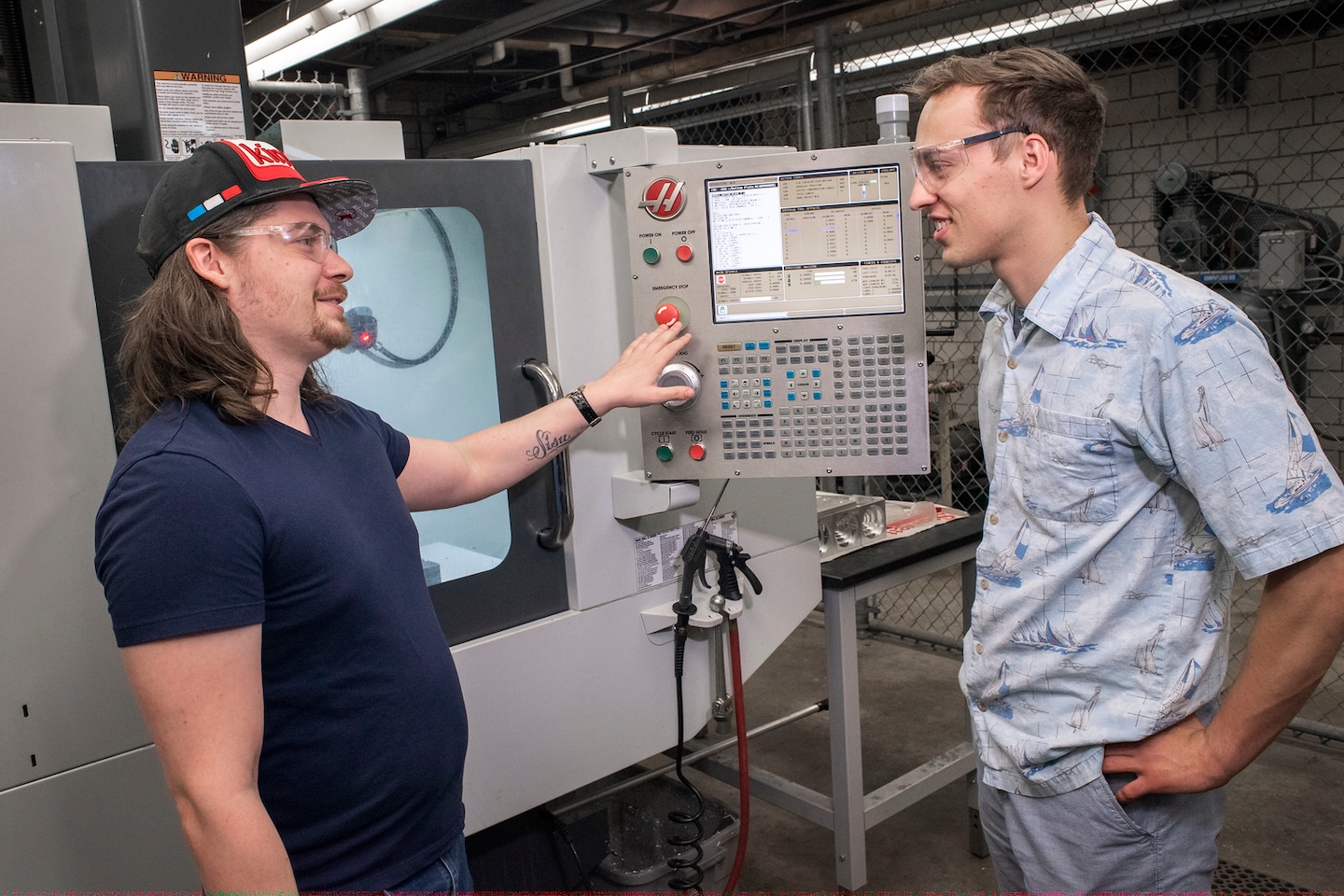Two young men are standing in a workshop, looking at each other and smiling while operating a Haas CNC machine. Both are wearing clear safety glasses.