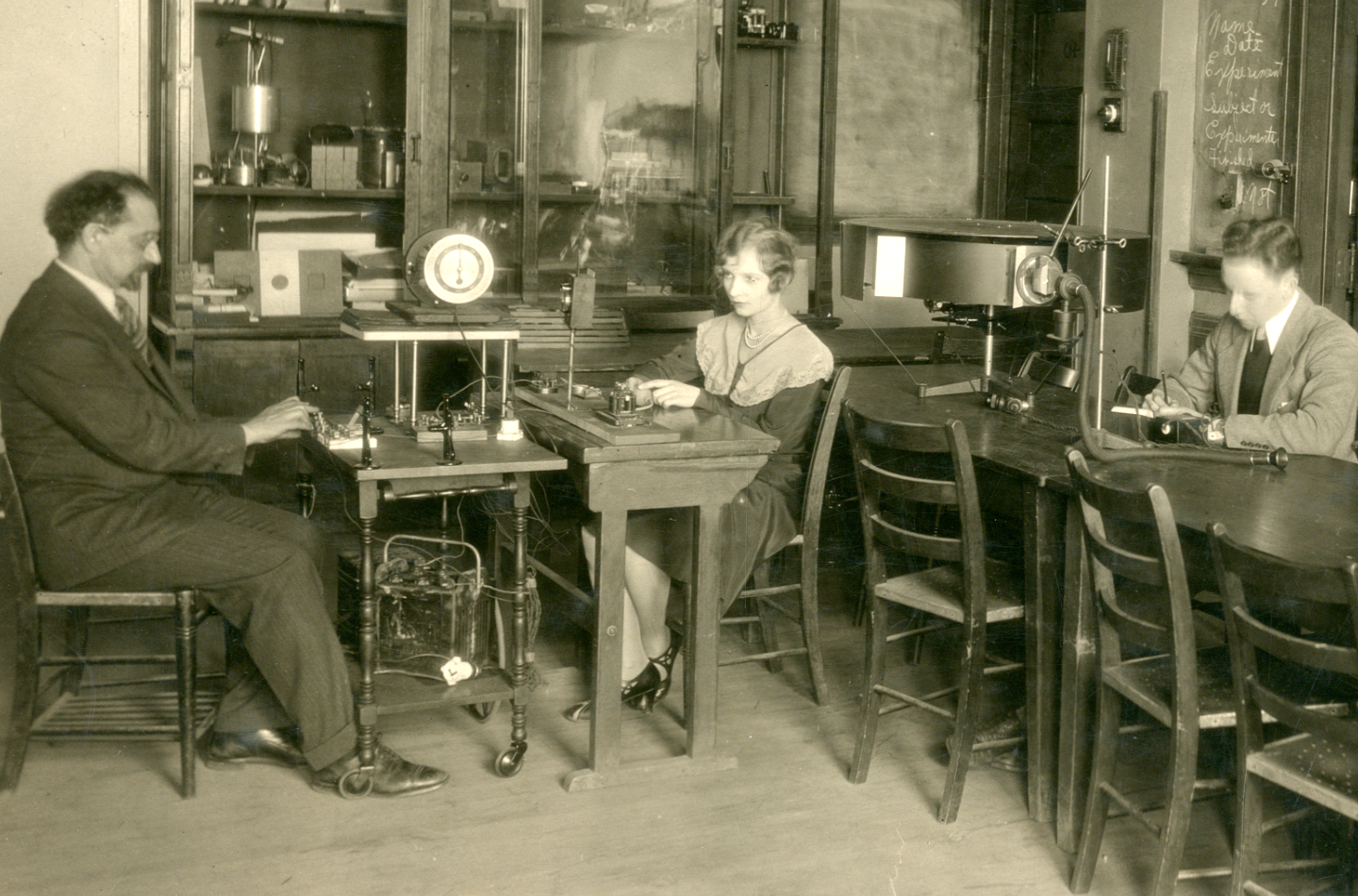 Three people sit at separate desks in a classroom or laboratory, working with scientific equipment and instruments, with shelves and a chalkboard in the background.