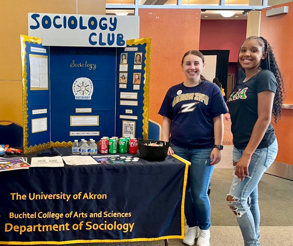 Two University of Akron students standing beside a Sociology Club information table and display board at a campus event.