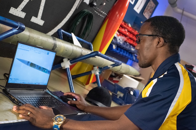 A person wearing glasses and a blue and yellow shirt works on a laptop displaying a 3D rocket model, with rocket parts and tools visible in the background.