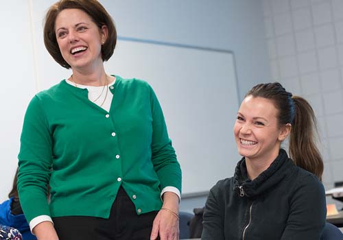 Two women smiling and laughing together in a classroom setting, one standing and one sitting, with a whiteboard in the background.