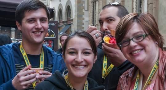 Four smiling college students wearing yellow lanyards enjoy cups of gelato on a cobblestone street during a study abroad trip in Europe.