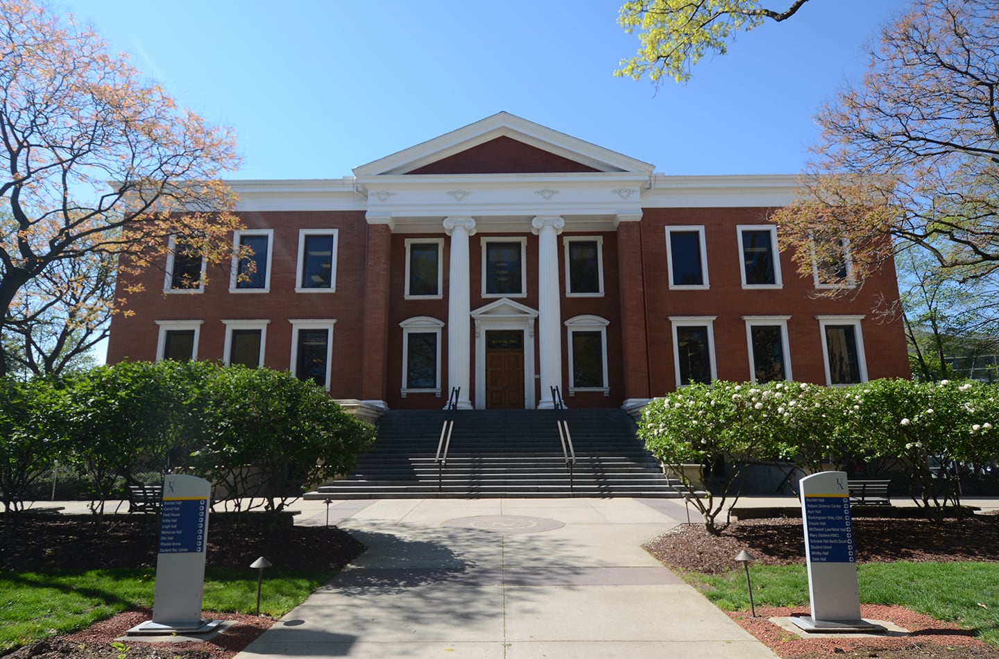 The front exterior of Buchtel Hall, a red brick building with white neoclassical columns and a pediment, featuring a large stone staircase and manicured campus landscaping.