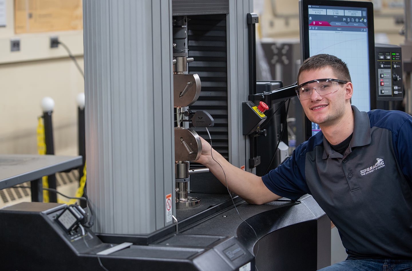 A female engineering student wearing safety glasses operates an Instron universal testing machine in a dimly lit laboratory. She focuses on a computer monitor displaying a real-time stress-strain graph as the machine performs a mechanical test on a small material specimen held in metal grips.