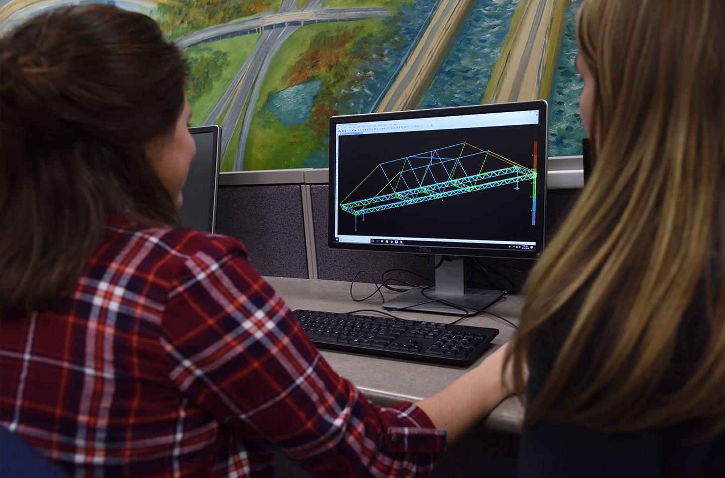 Two female engineering students working at a computer, viewing a 3D structural wireframe model of a bridge on a monitor. A painting of a highway interchange is visible on the wall in the background.