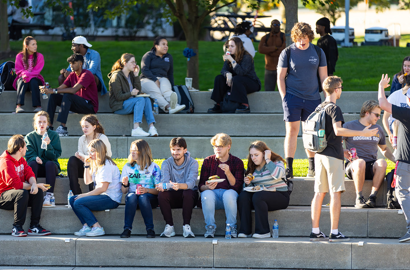 A group of college students gathering on large concrete outdoor steps on a sunny day. Many students are sitting and eating lunch or drinking from water bottles, while others stand and talk in small groups, illustrating social interaction on a university campus.
