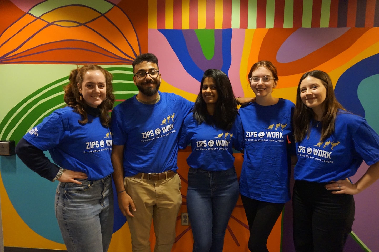 Five people stand side-by-side in front of a vibrant, multicolored abstract mural. They are all wearing matching bright blue t-shirts that read 