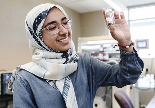 A woman wearing a patterned hijab and glasses smiles while holding up and examining a small vial in a laboratory setting.