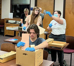Students wearing gloves examine archival materials from boxes during a hands-on class session in a library research room.