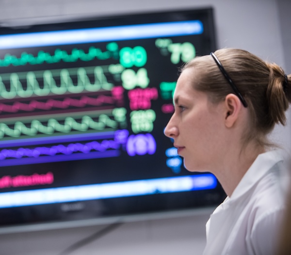 A woman in a white coat stands in front of a screen displaying colorful medical vital signs and heart rate graphs.