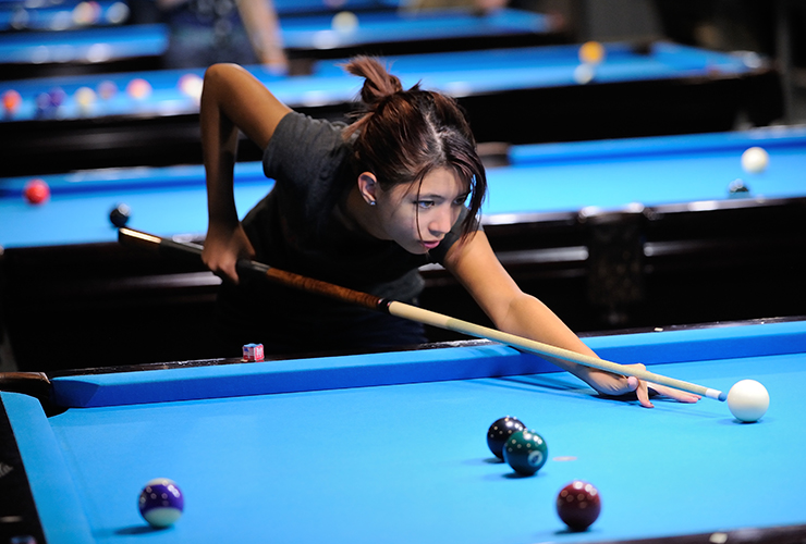 A young woman focuses intensely as she lines up a shot, aiming her cue stick at a white ball on a blue pool table. Several other colored billiard balls are scattered on the table.
