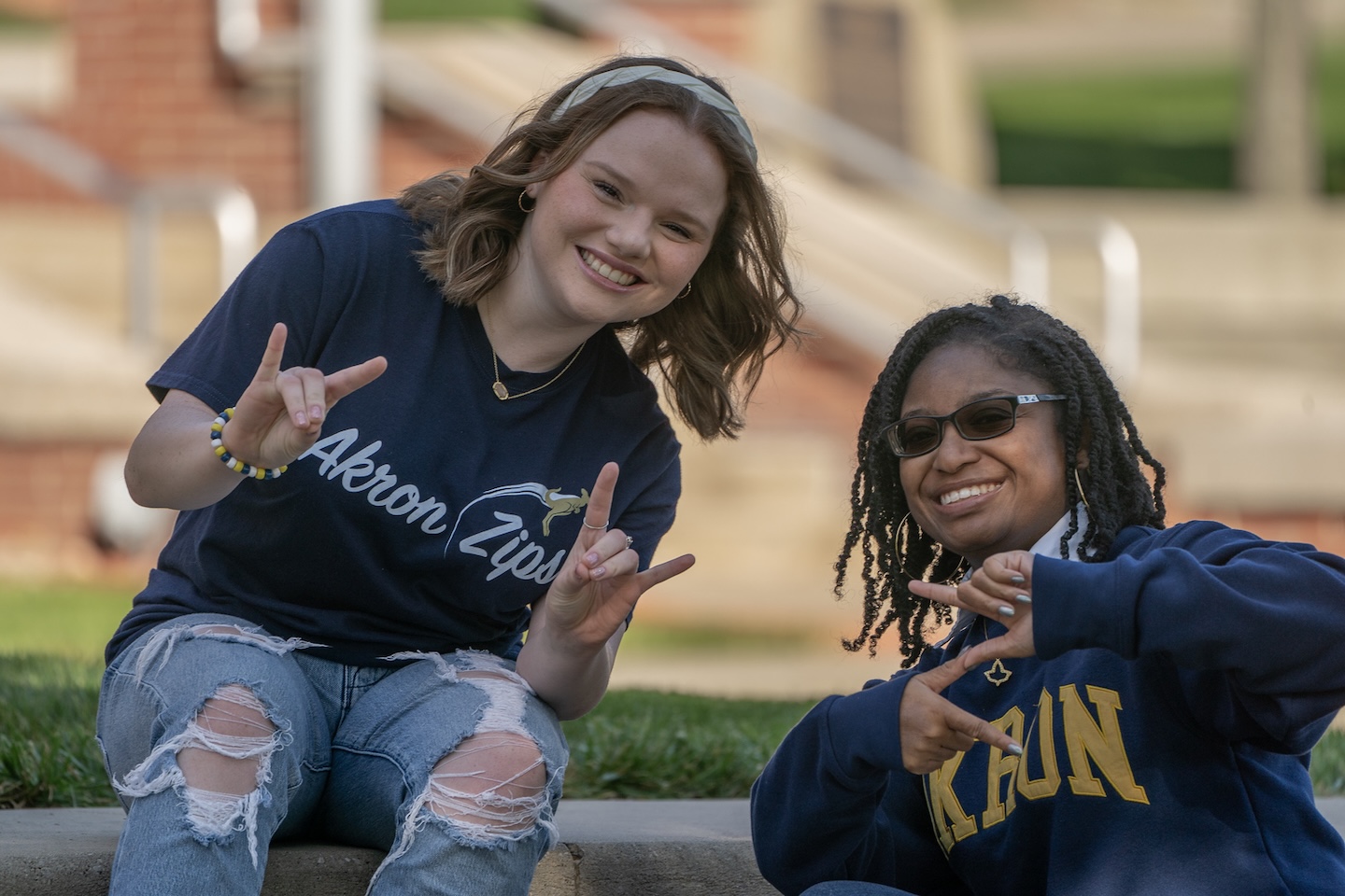 Two smiling University of Akron students sitting outdoors on campus, both making the Zips hand sign, wearing Akron Zips t-shirt and sweatshirt.