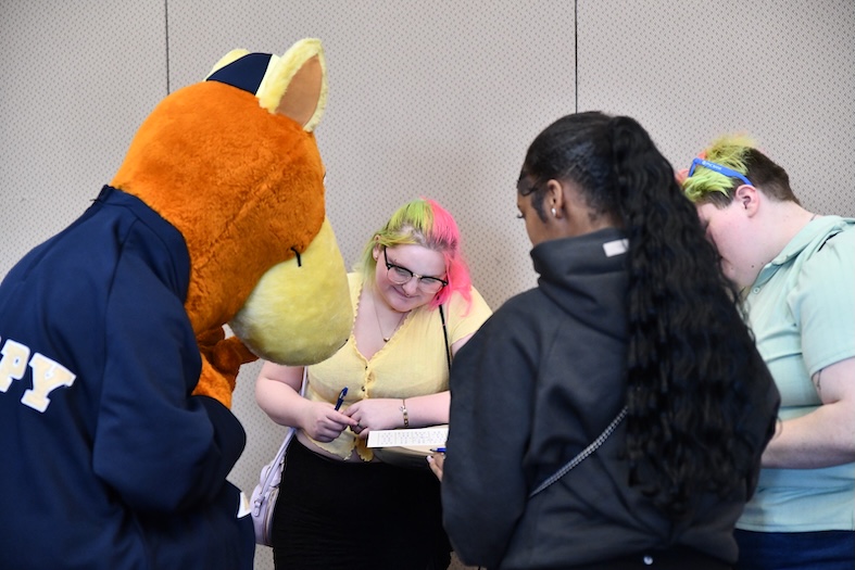 Students gather around a university mascot costume character, smiling and getting autographs during a campus event indoors.