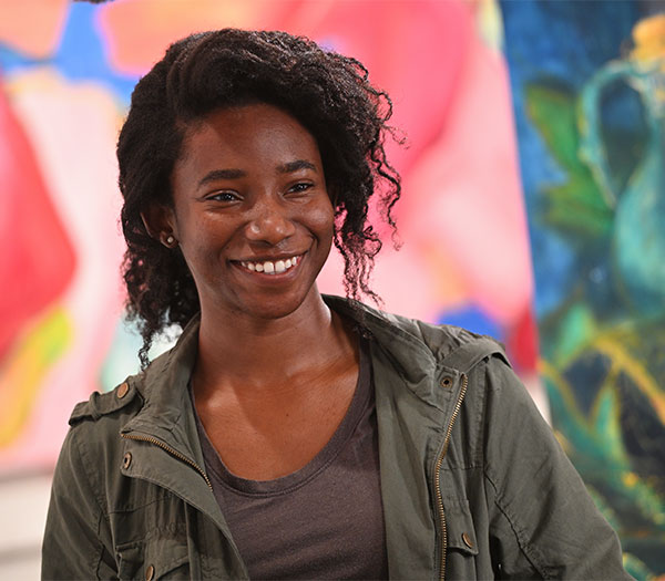 Art student smiles in front of colorful artwork displayed in a studio or gallery space.