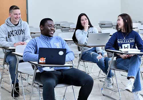 Four college students sit at desks in a classroom, smiling and talking together. Two have laptops open, and all appear relaxed and engaged. Empty desks fill the background.