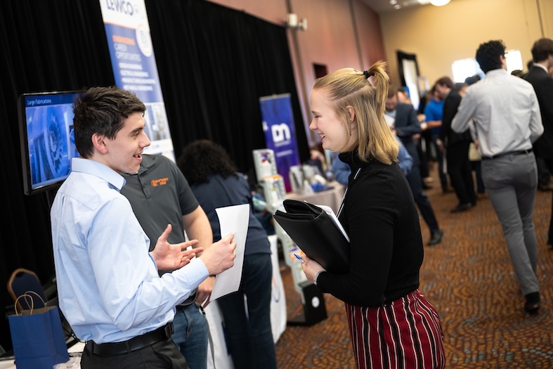 A student holding a portfolio folder speaks with a company representative at a university career fair, with employer booths and other attendees visible in the background.